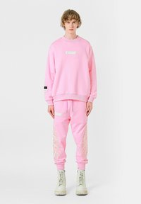 Sudadera rosa y joggers a juego con bolsillos laterales. La sudadera presenta una etiqueta blanca; los joggers tienen diseños de plumas bordados en blanco.