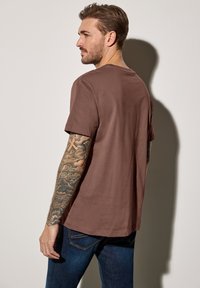 Hnědé bavlněné tričko s kulatým výstřihem a krátkými rukávy, nabízející volný střih. Zobrazuje hladkou texturu a šité detaily.