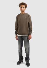 Bruine sweater met geribde manchetten, gecombineerd met vervaagde grijze jeans en zwarte sneakers. Bevat een klein wit logo op de borst.
