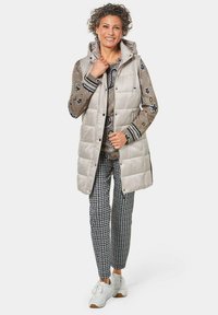 Wattierte Weste in hellbeige mit Kapuze, die ein gestepptes Design aufweist. Kombiniert mit einem gemusterten Strickoberteil und karierten Hosen. Weiße Sneaker.