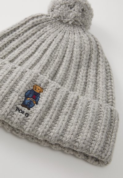 Grå ribbestrikket lue med en pom-pom på toppen. Har en brodert teddybjørn i en blå drakt og "Polo"-tekst på kanten.
