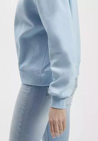 Jasnoniebieski sweatshirt z ryżowym wykończeniem przy mankietach i dole, miękka faktura oraz dopasowany krój, w połączeniu z jasnoniebieskimi dżinsami.