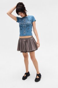 Blue short-sleeve t-shirt with ornate black pattern and rhinestones, paired with a gray pleated skirt and black footwear with cutouts.