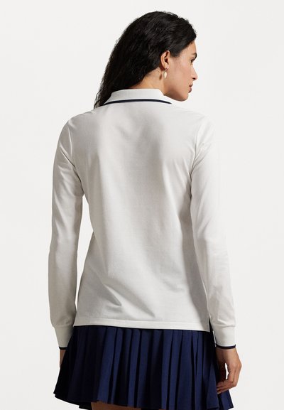 Witte polo met lange mouwen en marineblauwe kraag en zijaccenten, gecombineerd met een marineblauwe geplooide rok. Katoenen stof, gladde textuur.