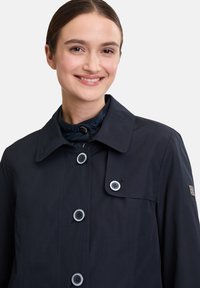 Granatowa kurtka przeciwdeszczowa z dużymi guzikami, szerokim kołnierzem i wewnętrznym pikowanym detalem kołnierza. Gładka tekstura i dopasowany krój.