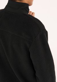 Pull noir en polaire avec col haut, surface texturée et accent sur la couture latérale. Présente un logo minimaliste embossé près de l'encolure.
