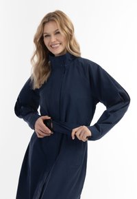 Laivansiniset tuulitakit, joissa on korkea kaulus, pitkät hihat, kiristetty vyötärö ja solki. Silkkinen pinta ja minimaalinen muotoilu.