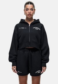 Felpa nera corta con zip e maniche oversize, abbinata a pantaloni corti neri coordinati. Presenta loghi bianchi e dettagli con cordino.