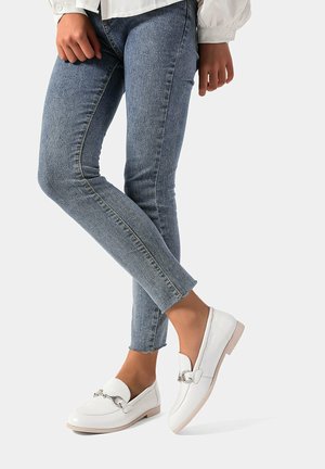 Mocassini in pelle bianca con un accento di catena argentata. Indossati con jeans attillati blu, evidenziando un look elegante e casual.