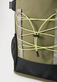 Zaino impermeabile verde con corpo testurizzato, base nera e cordini elastici verde lime a contrasto sul davanti per ulteriore spazio di stoccaggio.