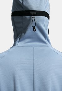 Ljust blå huvjacka med en texturerad tyg. Har en justerbar svart dragsko och släta, minimalistiska sömmar.