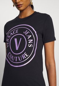 Černé bavlněné tričko s velkým, lesklým fialovým logem Versace Jeans Couture v kruhovém designu umístěným na přední straně. Krátké rukávy.