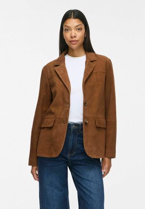 Vrouw draagt een bruine suède blazer over een wit overhemd met blauwe spijkerbroek, staand tegen een effen lichte achtergrond.