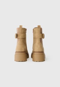 Bottines beige en suède, avec un talon bloc, une fermeture éclair à l'arrière, et des panneaux élastiques sur les côtés pour plus de flexibilité.