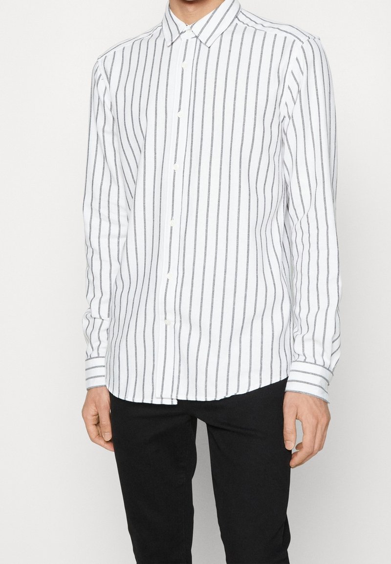 Camicia bianca a maniche lunghe con sottili righe verticali grigie, dotata di una chiusura frontale con bottoni, colletto a punta e polsini a righe contrastanti.