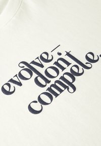 T-shirt en coton crème avec un grand texte en navy gras qui dit "évolue - ne te contente pas de rivaliser." avec une finition subtile en paillettes.