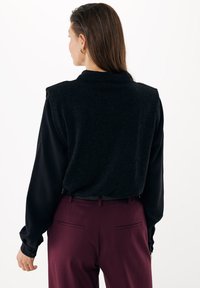 Černá mikina s vysokým límcem a ramenními vycpávkami, nošena přes košili s dlouhým rukávem. V kombinaci s vínovými upravenými kalhotami.