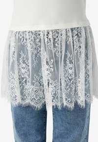 Jupon en dentelle florale blanche porté sur un jean en denim bleu et un top côtelé blanc, montrant un motif de dentelle complexe et une texture détaillée.