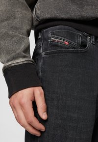 Black denim jeans featuring a slim fit, subtle fading, and a red and white logo tag. The cuff is contrasted with a textured, dark knit.