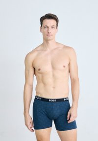 Shirtless man with short brown hair wearing blue patterned boxer briefs with black waistband labeled "BOSS" standing against light background.