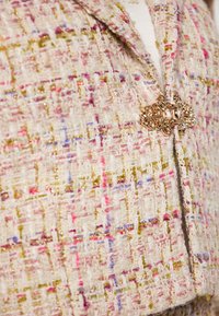 Blazer in tweed color crema, caratterizzato da fili colorati di rosa, viola e oro. Dettaglio di chiusura metallica decorativa sul davanti. Tessuto textures.