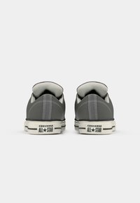 Pelēki auduma zemi sneakeri ar baltām gumijas zolēm, kas izceļas ar "CONVERSE ALL STAR" zīmolu un kontrastējošiem šuvumiem aizmugurē.