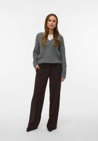 Szary sweter z wełny z dekoltem w serek i ściągaczami na rękawach, zestawiony z ciemnobrązowymi, szerokimi spodniami. Modelka stoi z rękami w kieszeniach.
