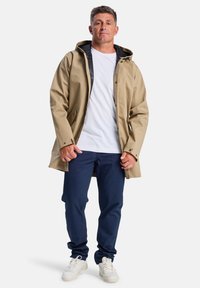 Un homme se tient avec assurance, portant un imperméable beige sur une chemise blanche, un pantalon bleu marine et des baskets blanches, avec un arrière-plan neutre.