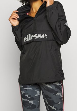 Osoba nosząca czarną kurtkę typu windbreaker marki ellesse i szare legginsy w kamuflażu z różowym paskiem, poprawia kołnierz kurtki.