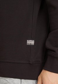 Black sweatshirt with ribbed texture, featuring a small white G-Star RAW logo tag at the side seam. Simple design, no patterns or accents.