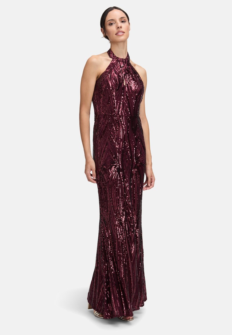 Frau in einem ärmellosen, bodenlangen, mit Pailletten besetzten weinroten Kleid mit Halterneck, stehend vor einem einfachen, hellen Hintergrund.