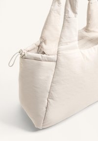 Lys beige tote taske lavet af blødt stof. Har to håndtag, en lynlås lukning og en samlet top med en snøre detalje.