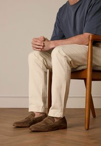 Bruine suède instappers met een ronde neus en gestikte details, gecombineerd met beige slim-fit broeken, zittend op een houten stoel.