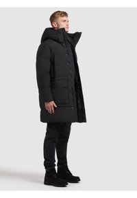 Cappotto imbottito nero con colletto alto, zip frontale e bottoni a pressione. Presenta grandi tasche anteriori e una texture liscia. Indossato sopra jeans scuri e stivali.