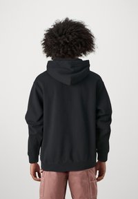 Sudadera negra con una gran capucha con cordón, puños y dobladillo acanalados, hecha de un tejido suave. Llevada con pantalones cargo rosas claros.
