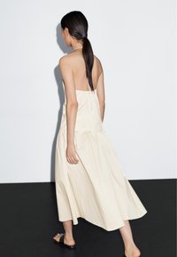 Woman with long dark ponytail walking barefoot on black floor wearing a backless, cream-colored halter dress with black slide sandals.