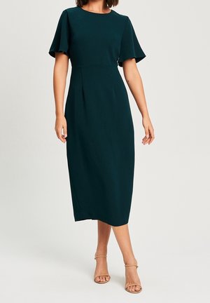 Frau trägt ein dunkelgrünes Midikleid mit flatternden Ärmeln und beigefarbene, geschlossene High-Heel-Sandalen mit Riemchen, steht vor einem einfarbigen Hintergrund.