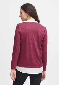 Maglietta a maniche lunghe color marrone con una consistenza morbida, caratterizzata da un collo a giro e un look a strati con una camicia bianca all'orlo.