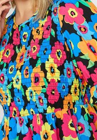 Lähikuva monivärisestä kukkakuosisesta mekosta, jossa on kootun bodyn ja puffihihat kirkkaan vaaleanpunaisena, sinisenä, keltaisena, oranssina ja vihreänä mustalla taustalla.