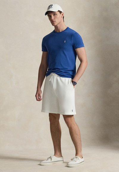 Μπλε εφαρμοστή μπλούζα, λευκό σορτς με κορδόνι, λευκά αθλητικά παπούτσια και λευκό καπέλο με λογότυπο, όρθιος σε ουδέτερο φόντο.