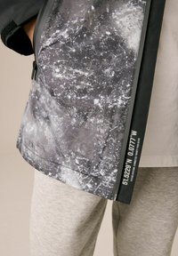 Giacca stampata in grigio e nero con superficie texture e chiusura con zip. Include coordinate e dettagli in colori a contrasto.