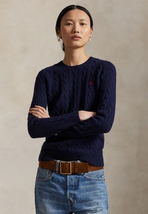 Sötét hajú nő, aki tengerészkék, fonott mintás pulóvert, barna velúr övet és világoskék farmert visel, karját keresztbe téve áll semleges háttér előtt.