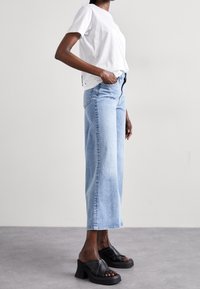 Camiseta blanca de algodón, corte relajado; jeans rectos azul claro; sandalias negras de tacón con tiras entrecruzadas. Vista de perfil lateral.