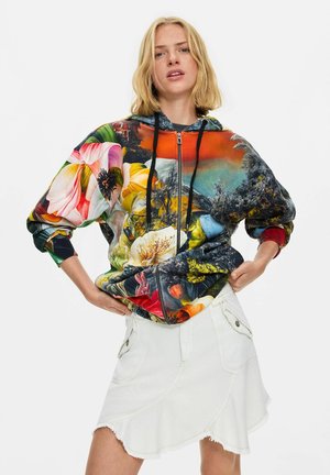 Junge Frau trägt einen bunten floralen Reißverschluss-Hoodie mit schwarzen Kordeln und einem weißen asymmetrischen Rock mit Taschen, steht vor einem schlichten Hintergrund.