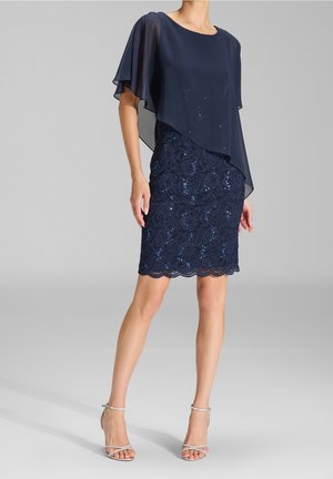 Frau in einem marineblauen Spitzenkleid mit Pailletten bis zum Knie, mit einem transparenten asymmetrischen Cape und silbernen Riemchen-High Heels.