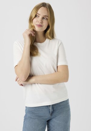 Balta T-krekls ar apaļu kakla izgriezumu, īsām piedurknēm un brīvu piegriezumu, izgatavots no gludas kokvilnas auduma. Kombinēts ar zilām džinsa biksēm.