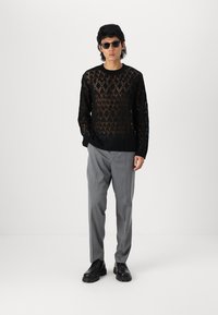 Svart åpenstrikket genser med diamantmønster, kombinert med grå bukser. Modellen har på seg mørke solbriller og svarte sko. Minimalistisk design.