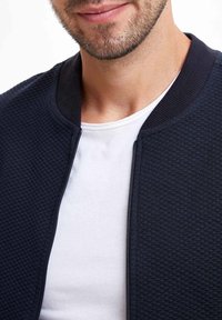 Granatowa pikowana kurtka zapinana na zamek z prążkowanym kołnierzem, noszona na gładkim białym t-shircie, prezentująca teksturowany wzór tkaniny.