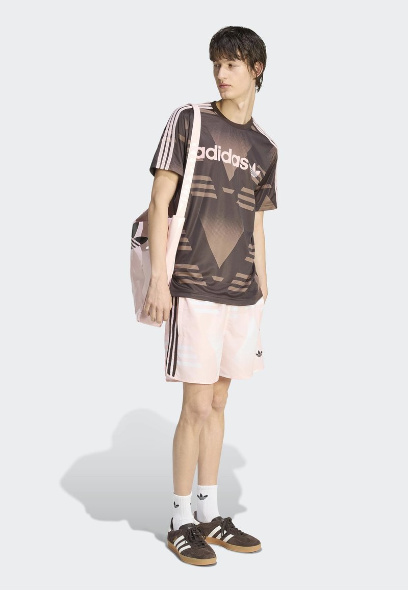 Giovane uomo che indossa una T-shirt Adidas scura con motivo a fantasia, pantaloncini Adidas rosa chiaro, calzini bianchi Adidas e scarpe da ginnastica Adidas marroni, mentre porta una borsa a tracolla rosa.