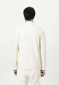 Geripptes, elfenbeinfarbenes Sweatshirt mit langen Ärmeln, hohem Kragen und schwarz gestreiften Bündchen. Weiche Textur und lockere Passform sind von der Rückansicht deutlich zu erkennen.